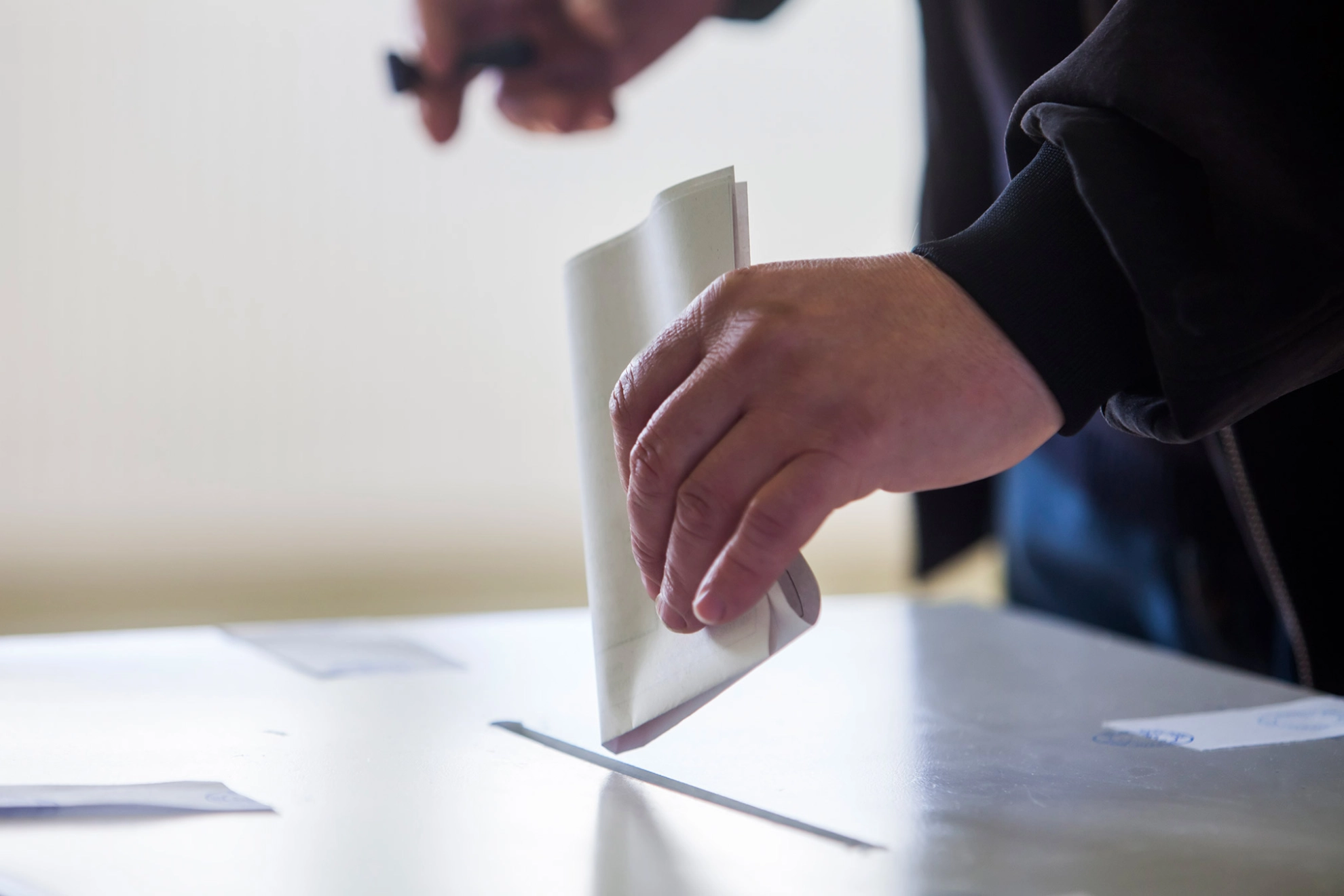 Jemand steckt mit der linken Hand einen gefalteten Wahlzettel in ein Wahlurne.