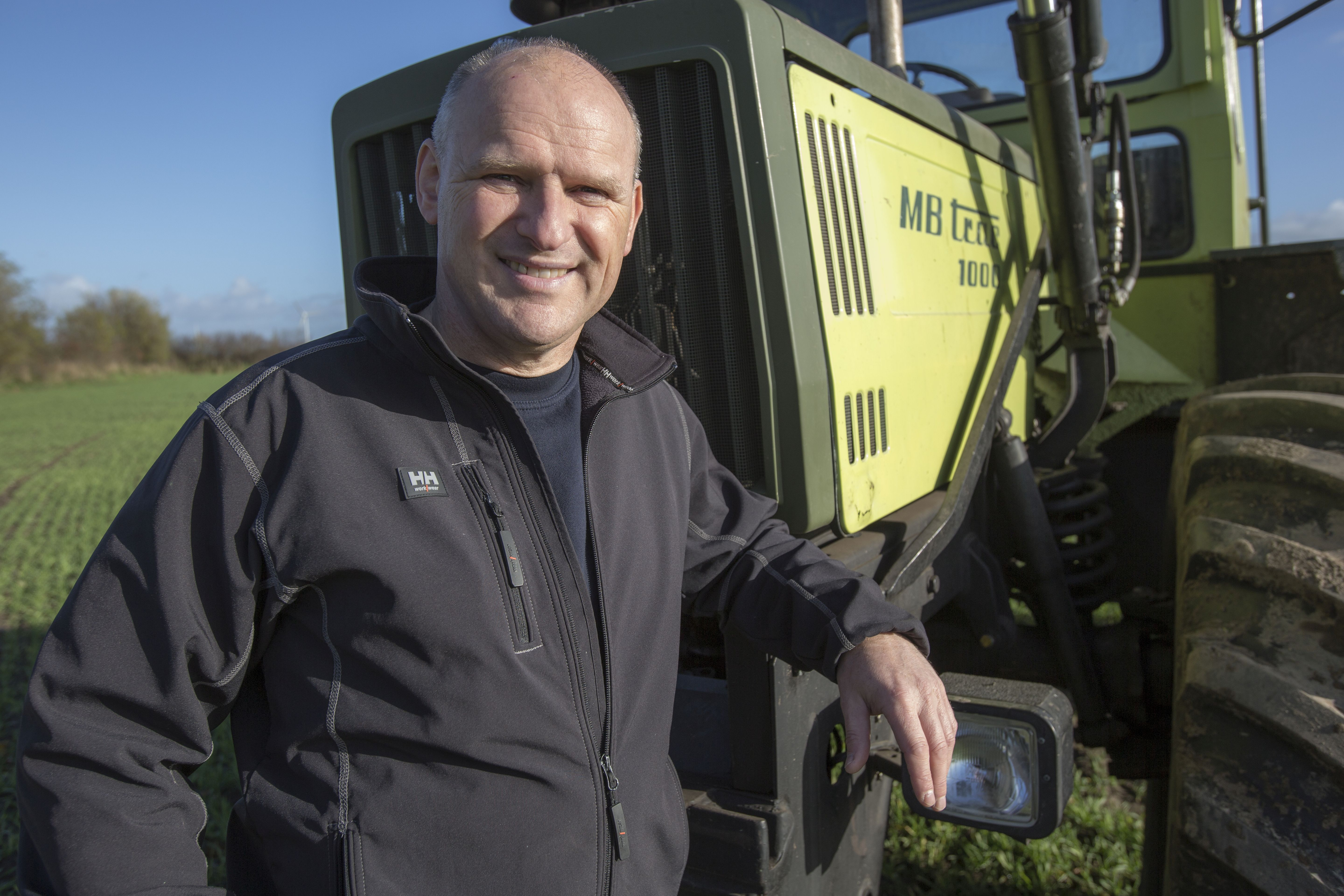 Vestas Monteur Arno Buß steht auf einem schleswig-holsteinischen Acker und lehnt an einem gelben Traktor.